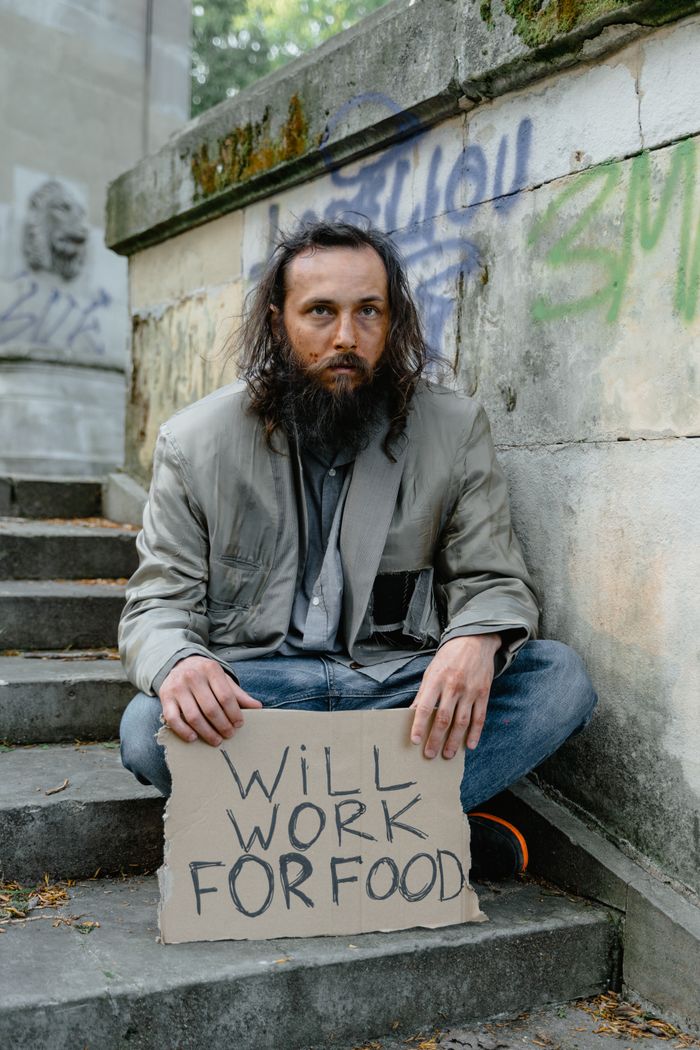 Homeless man with long hair and beard holding 'Will work for food' sign on graffiti-covered stairs.